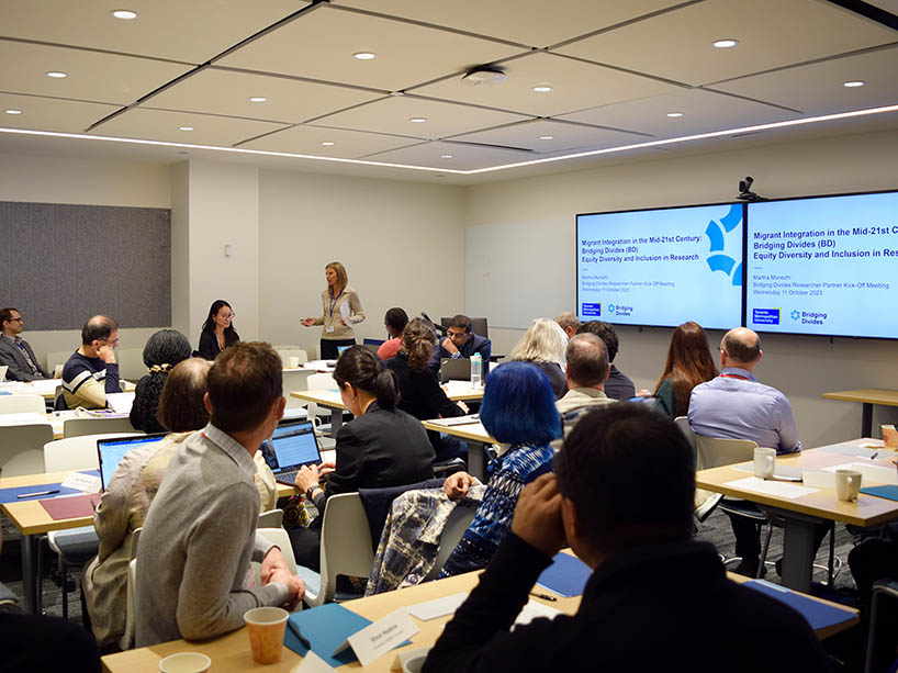 A group of people working together in a room are listening to a speaker in front of two screens.