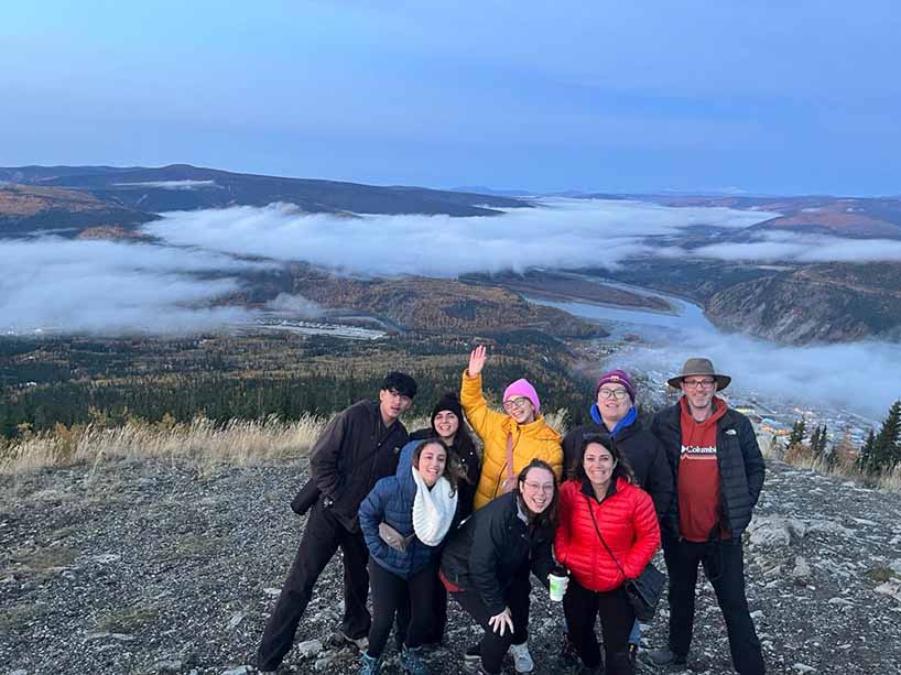 A group of students pose and wave in front of a valley with rivers and clouds.