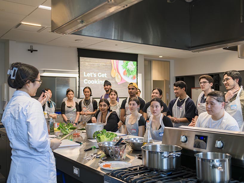 A student holds a microphone in front of cooking materials while other students look on