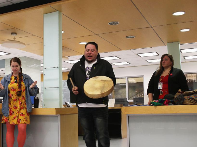 Wayne Moberly sings with his hand drum, community members stand beside him. 