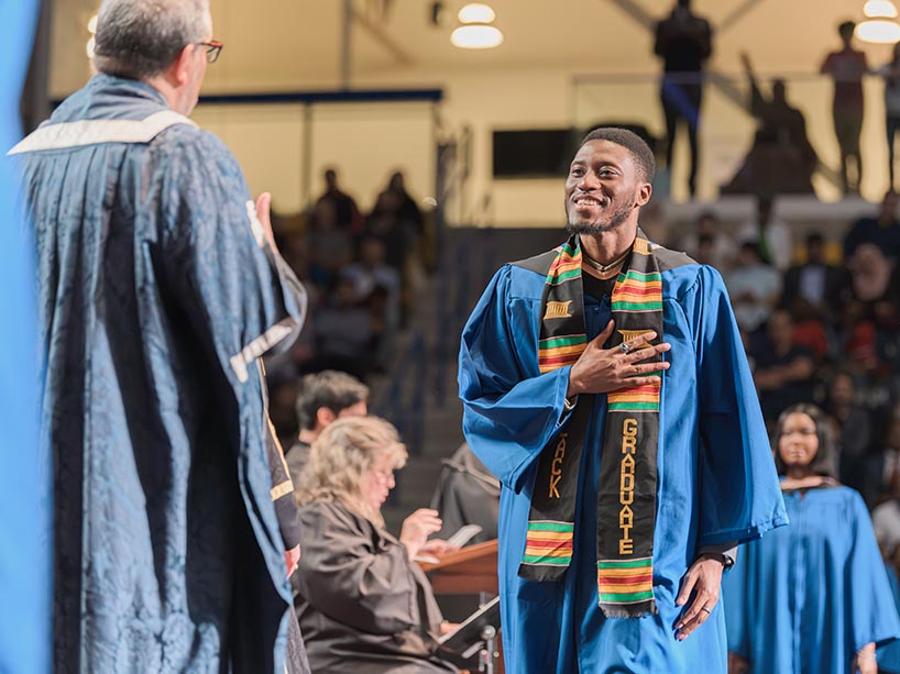 A Black student crosses the stage at convocation. He is wearing a blue gown with a traditional Kente stole around his neck.