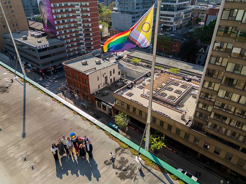 An aerial view of a group of people on a rooftop with a large flag pole. There are other tall buildings surrounding them.