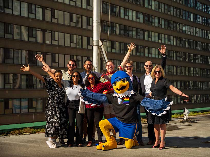  A group of people and a falcon mascot stand on the rooftop of a building, posing with a large flag pole with their arms stretched out