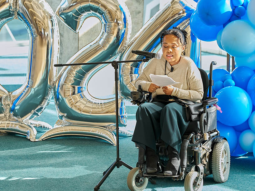 Kate Tutu on a mobility aid device, speaking into a microphone with a backdrop of balloons.