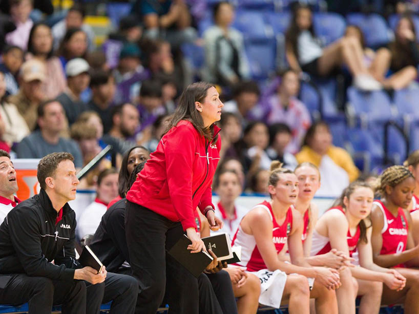 A female coach in a red top stands by a team bench, looking out toward the basketball court.