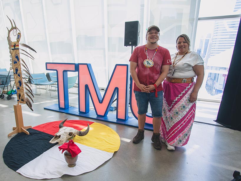Waasekom and Crystal Osawamick stand in front of the TMU letter with the TMU eagle staff, buffalo skull and medicine wheel blanket.