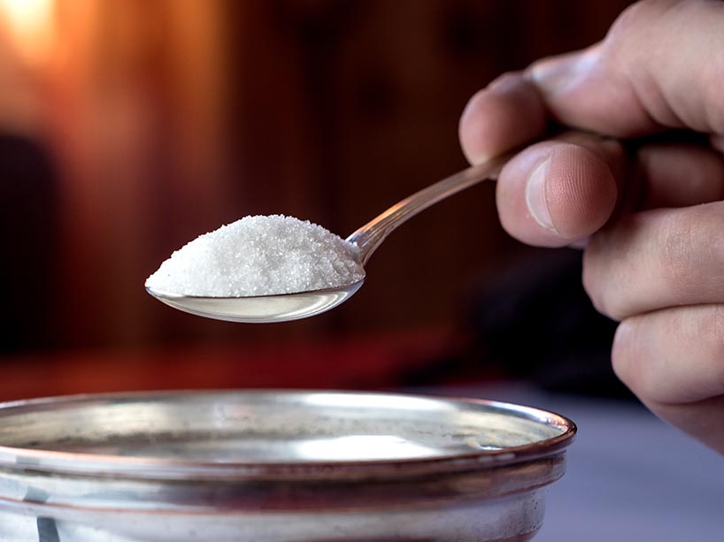 A hand holds a teaspoon of sugar over a jar.