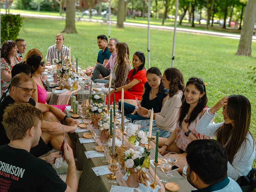 A group of people sit together at picnic tables in a park. The tables are decorated with candles and flowers.