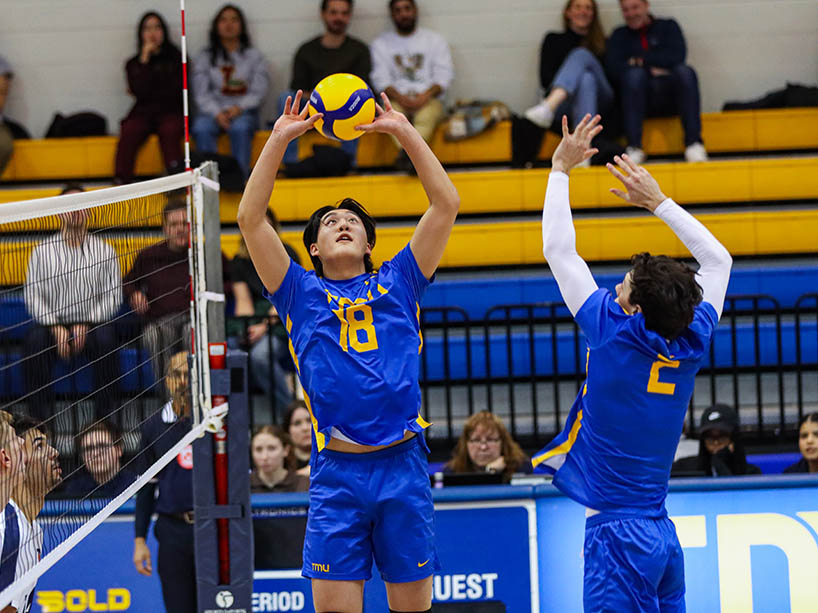 A TMU Bold player begins to set the ball for a teammate, who prepares to spike it over the net.