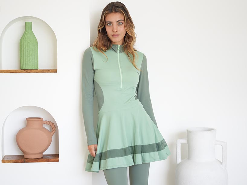 Young woman wears a light green full-coverage swimsuit as she leans against a wall shelf with colourful vases