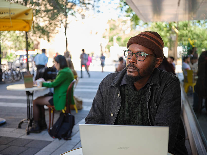 Victor Tubuseeke sits at an outdoor patio on campus with his laptop.