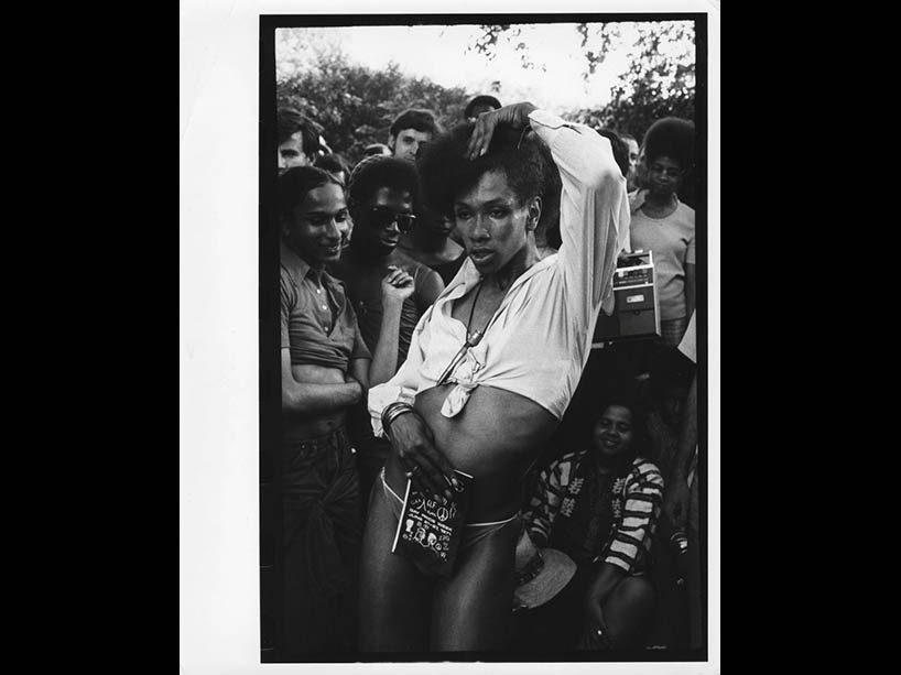 Feminine-presenting BIPOC dancer with a tied cropped top strikes a pose while holding a pamphlet in front of a crowd of onlookers from the 2SLGBTQ+ community.