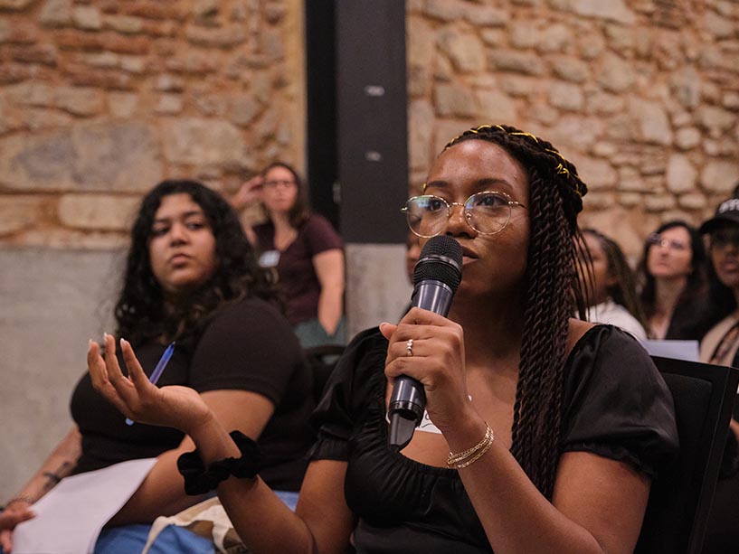 A Black student speaks into a microphone at a TMU event, while other students listen nearby.