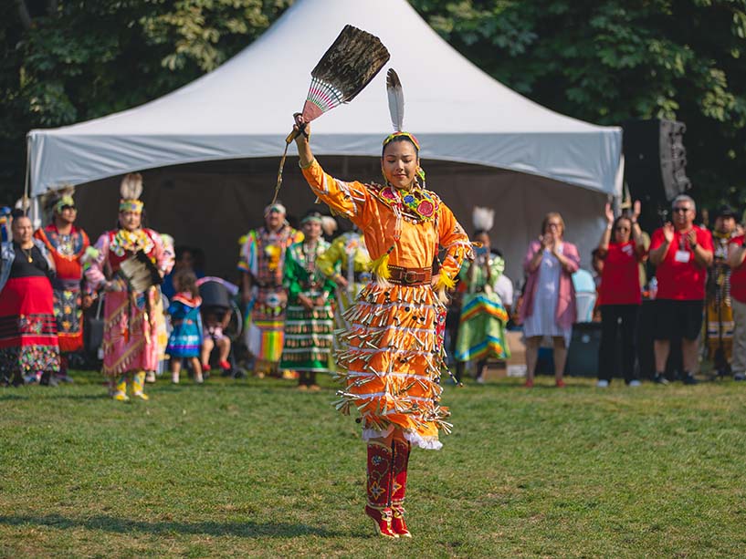 In her orange jingle dress, Madison Noon dances in the Pow Wow circle with her eagle fan raised above her head.