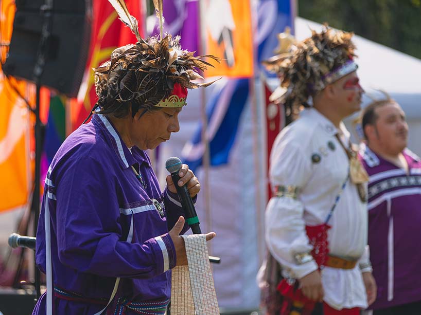 Wearing a purple shirt with white ribbons and a headpiece adorned with mostly brown feathers, Frank McNaughton holds a wampum belt in his right hand as he speaks into the microphone in his left.