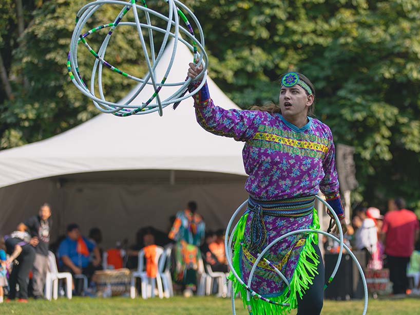 A hoop dancer in purple and green regalia tosses several hoops in a sphere formation in their right hand while balancing other hoops around their hips and waist.