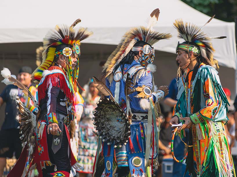 Three dancers — two are wearing regalia with feathered bustles on their backs and one is wearing fringed regalia — speak amongst themselves between dances.