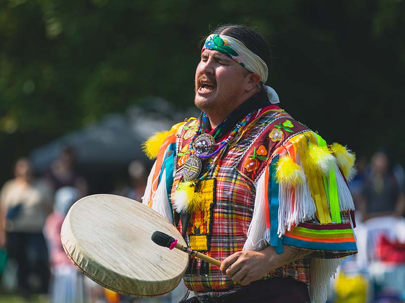 A drummer in colorful regalia adorned with shoulder fringes and small yellow feathers sings and drums with their eyes closed. 