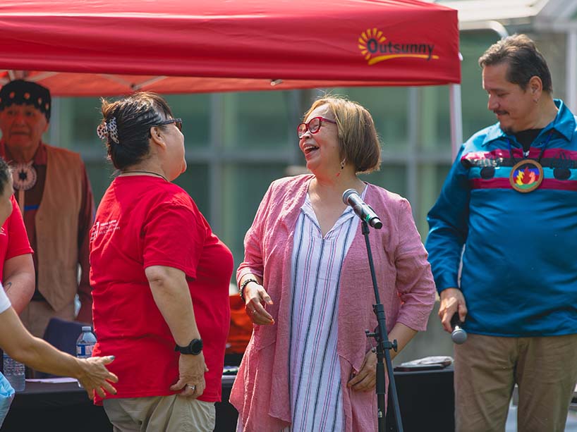 Monica Mckay, dressed in pink and white, and Sheila Saikkonnen, wearing a red t-shirt, laugh together next to the microphone. Behind them dressed in a blue shirt, Bob Goulais, Pow Wow MC, smiles. 