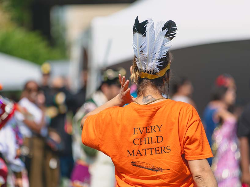 A Pow Wow attendee faces away from the camera. They are wearing a headband with feathers and an orange shirt which reads, “Every child matters.”