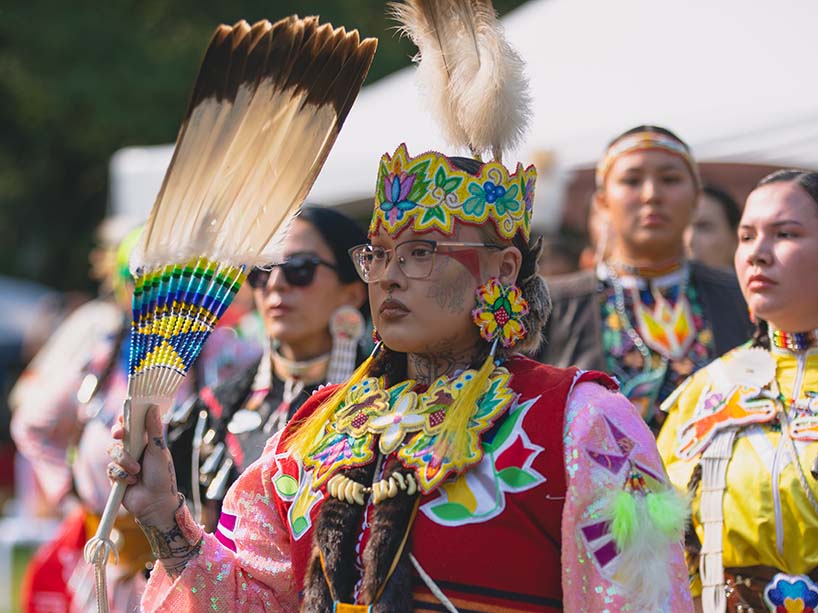 Eagle fan held aloft, dancer Aj Douglas, whose regalia includes a yellow beaded headband with an eagle feather and beaded earrings with a long yellow fringe, waits to dance.