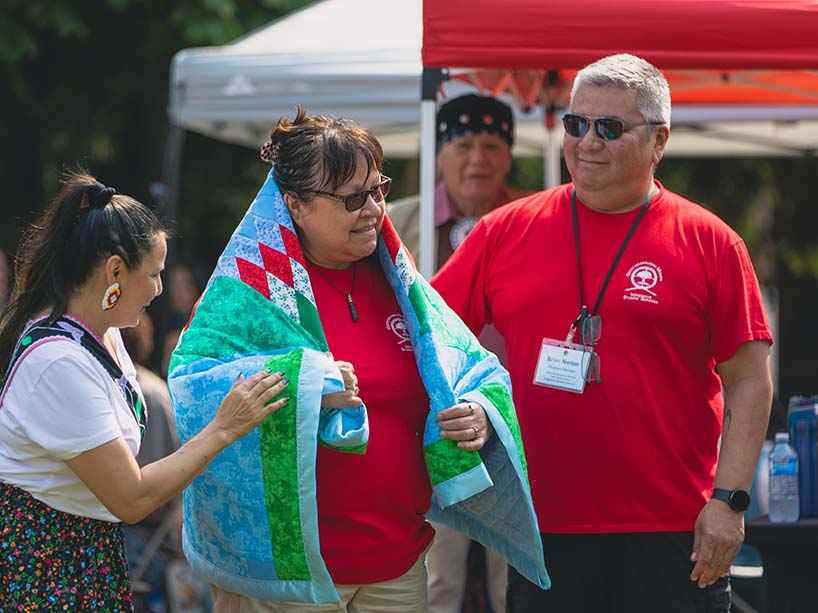 Sheila Saikkonen wraps her newly gifted blanket, which is green, blue, white and red around her shoulders. On either side of her, Cher Trudeau, dressed in a white t-shirt and floral ribbon skirt, and Brian Norton, wearing a red t-shirt, smile.