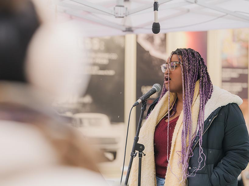 A young woman with long braids, wearing a winter coat and scarf, speaks into a microphone at an outdoor winter event.