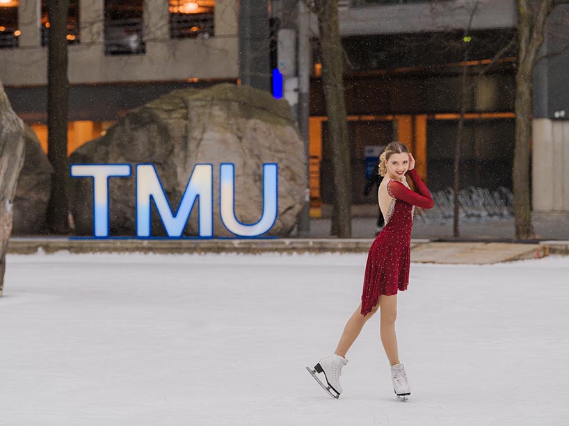 A figure skater in a red dress and white figure skates poses on the ice rink at TMU.