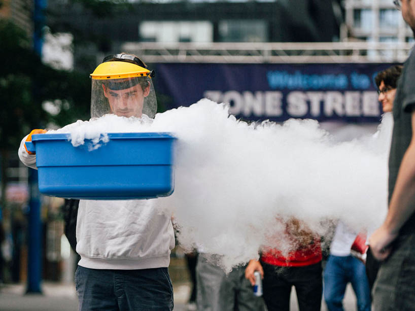 A man wearing a face shield and gloves holds up a plastic box. The box has white smoke coming out of it.