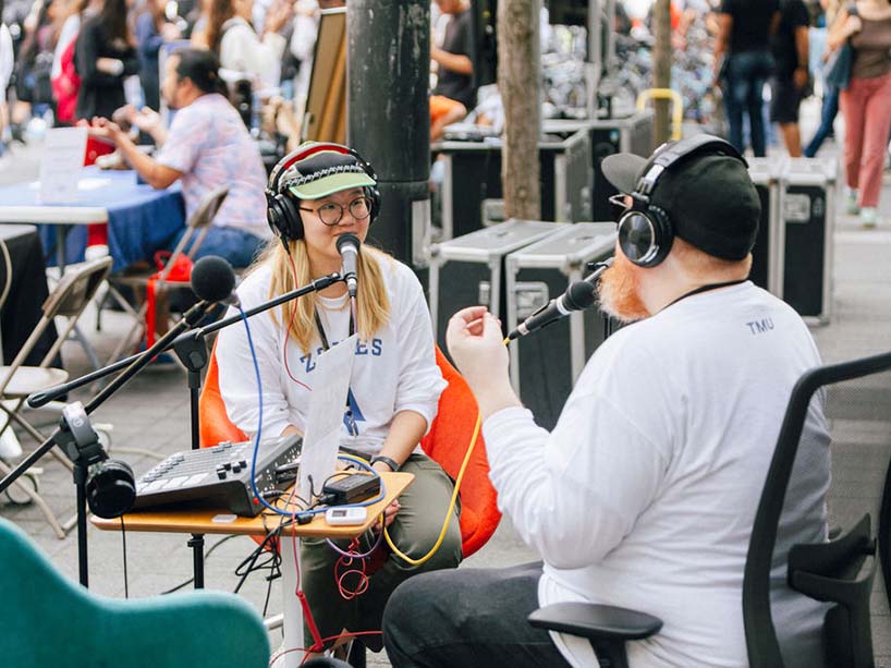 Two people wearing headphones sit at a table and talk into microphones.