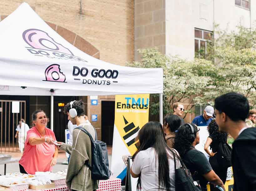 A crowd of people line up at a tent that reads “Do Good Donuts” while a woman hands out baked goods.