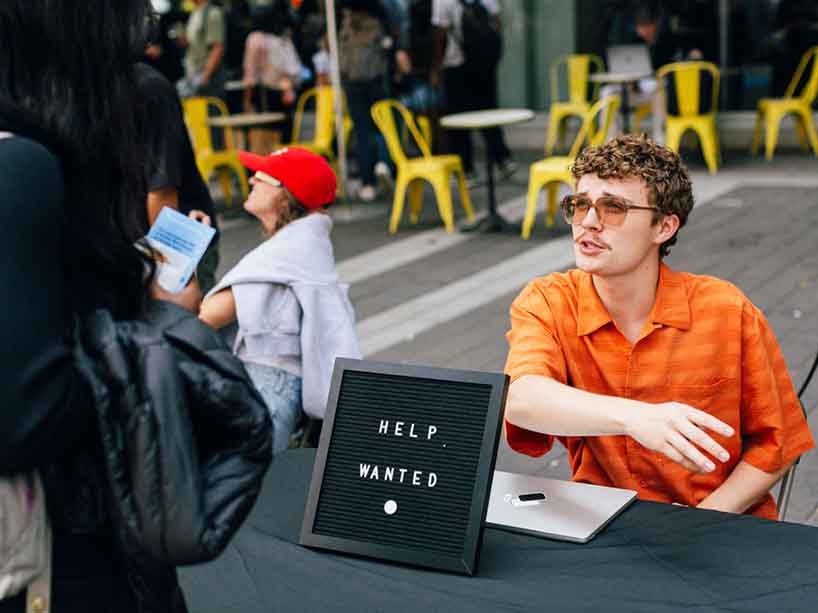 A man sitting at a table with a “Help Wanted” sign talks to a student.