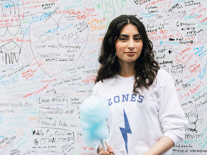 A person wearing a shirt that says “Zones” stands in front of a white board with writing of startup ideas and sketches on it.