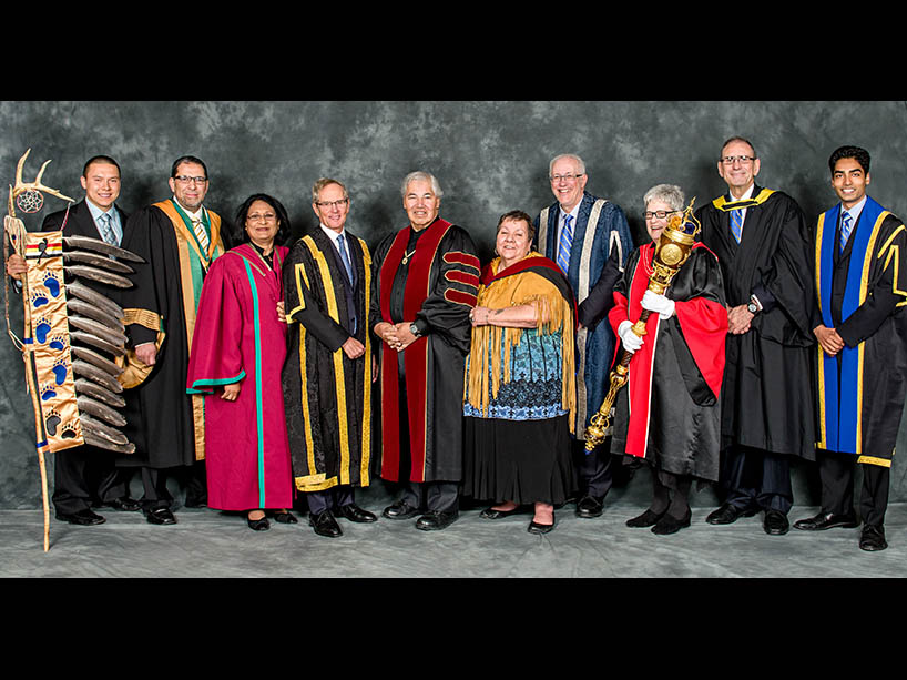 Murray Sinclair, wearing graduation robes stands with university dignitaries in robes. One person holds the Eagle Staff while another holds the convocation mace.