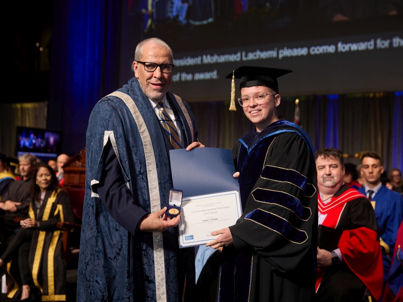 Two people wearing academic regalia pose for a photo while holding a certificate and a gold medal.