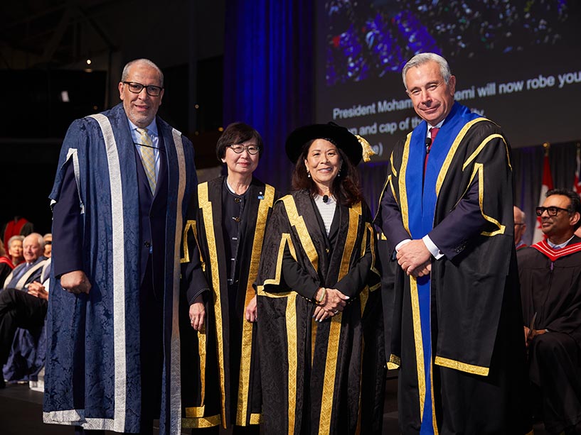 Four people pose for a photo on a stage. They are wearing academic regalia. 