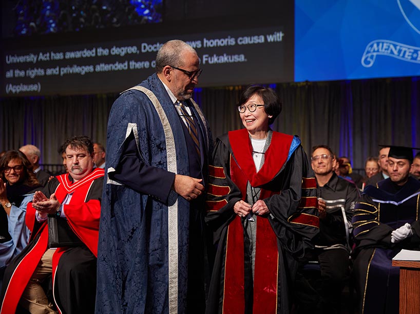 A man and a woman wearing academic regalia stand together and smile at each other during a ceremony.