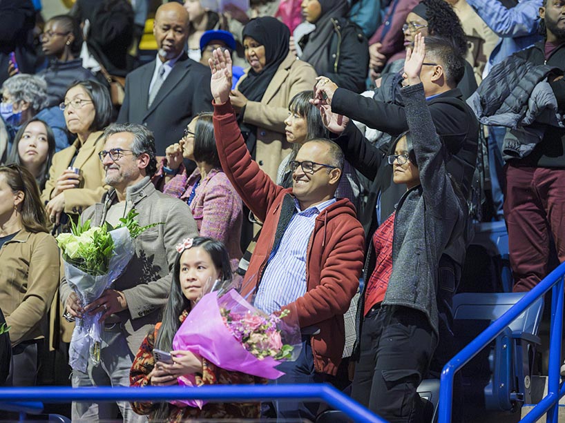 A crowd of people in stands watch a ceremony. Some wave while others take photos or hold flowers.