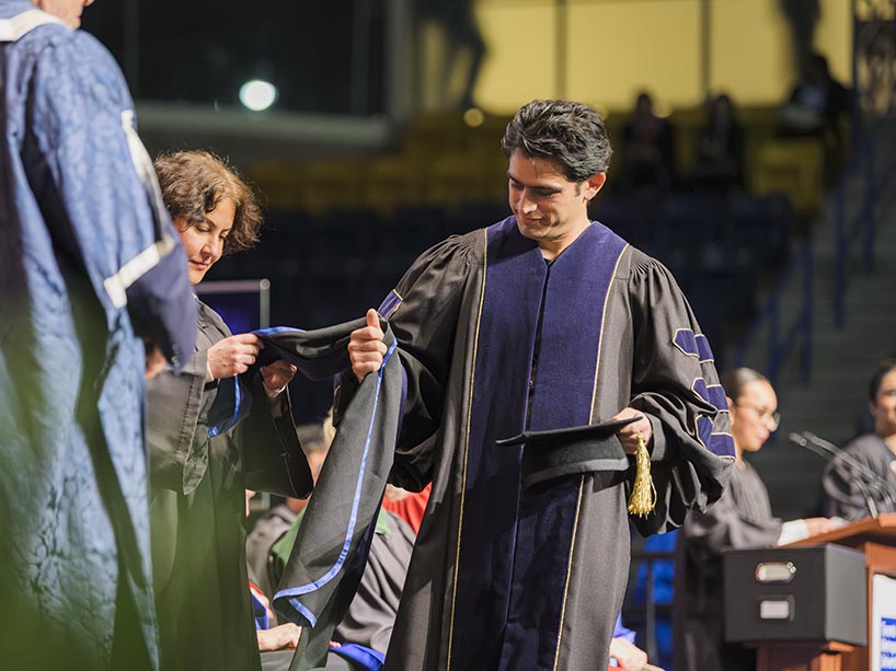 A young man walks across a stage holding a hood/scarf and mortarboard hat.