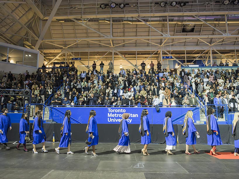 Students walk in a line. They are walking in front of crowds of people sitting in stands.