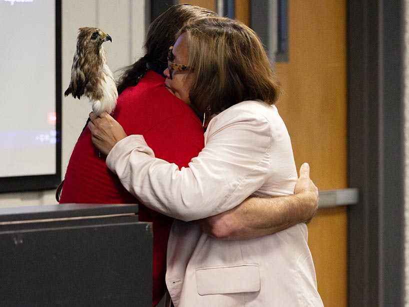 Wearing a red t-shirt, Bradley Gibson, Indigenous Academic Student Advisor, hugs McKay, wearing a pale pink blazer and holding a preserved hawk in her left hand. 