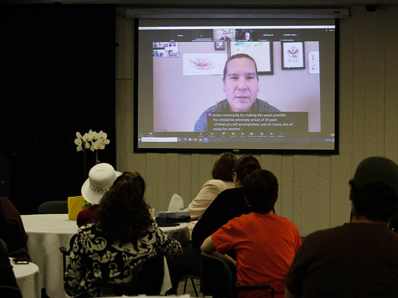 Waubgeshig Rice appears virtually to deliver his keynote speech from his home office. Artwork by his children hangs on the wall behind him. Event attendees, including McKay and her mother, Audrey McKay, attentively watch the screen. 