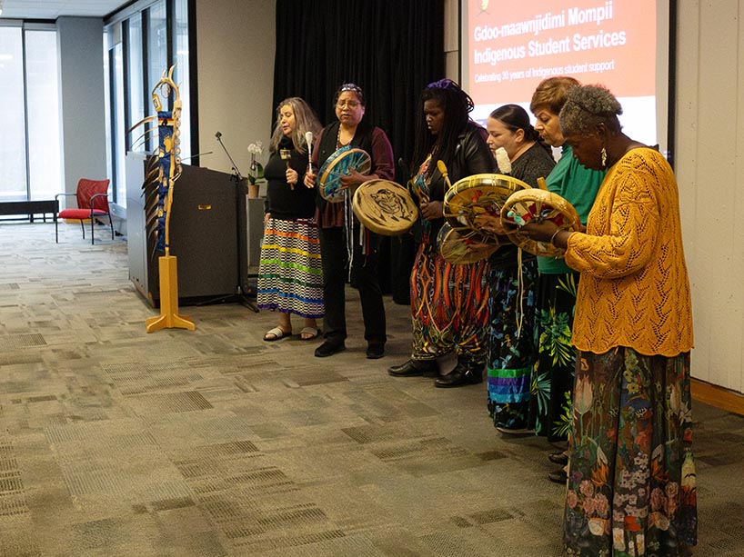 Six members of the Spirit Winds drum group stand at the front of the room and perform an honour song. Some members are wearing ankle-length ribbon skirts.