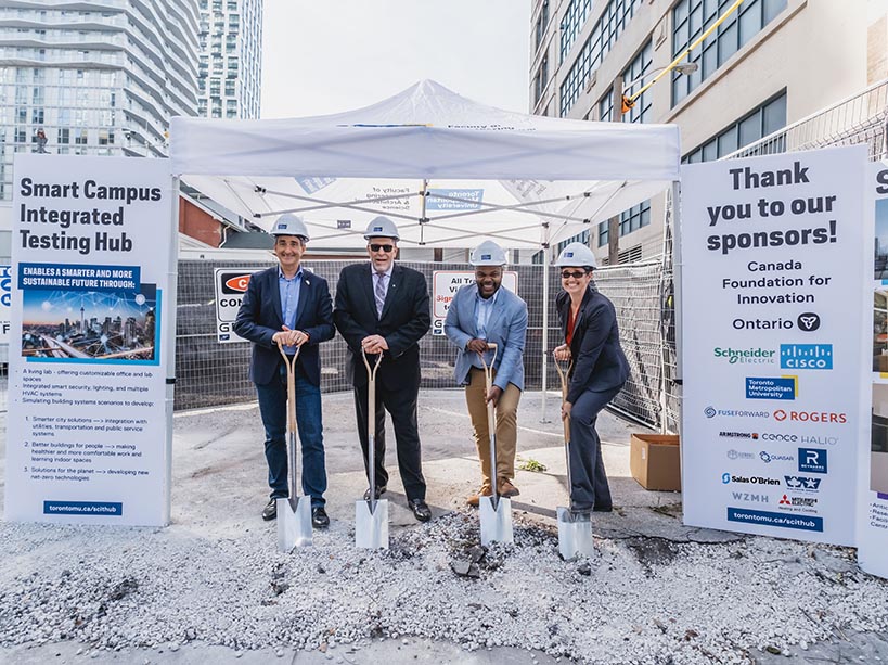 Four people wear hard hats and hold shovels on a construction site