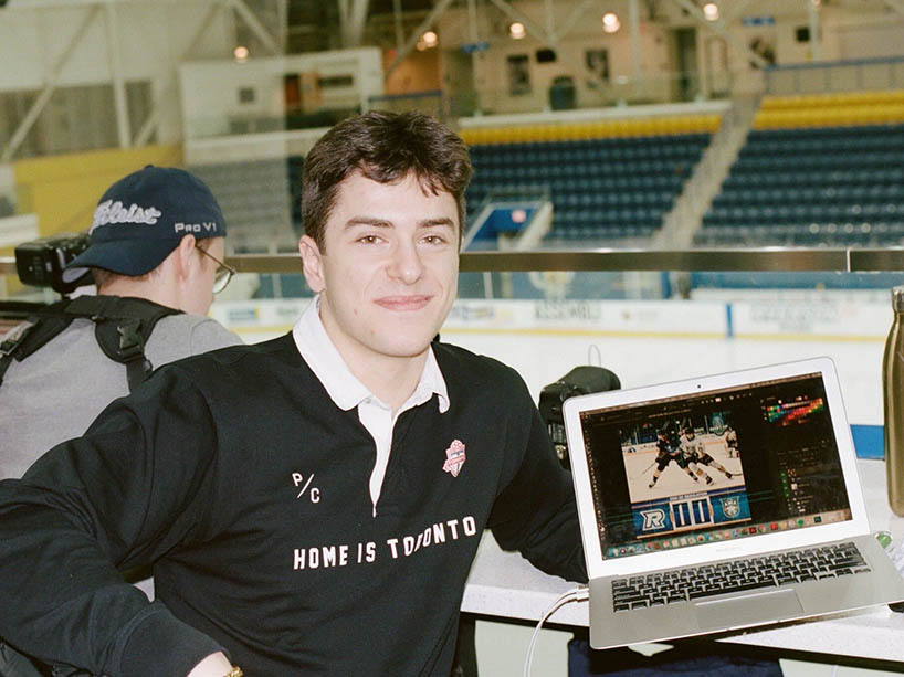 Émile Riga smiles with a hockey game shown on his laptop, and a hockey rink behind him.