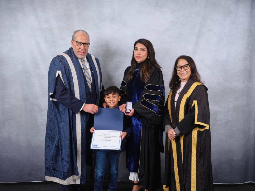 Three adults wearing academic regalia and a child pose for a photo while holding a certificate and a gold medal.