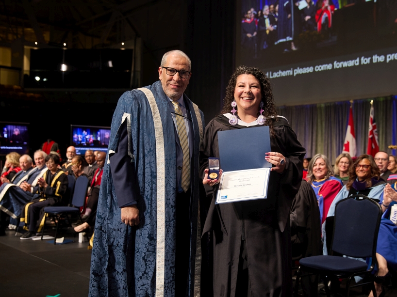 Two people wearing academic regalia pose for a photo. One person is holding a certificate and a gold medal.