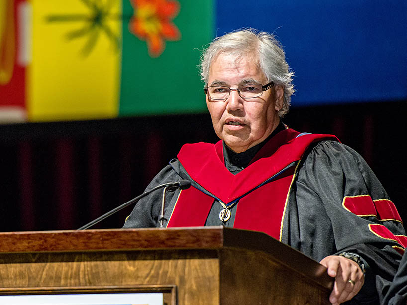 Murray Sinclair, wearing graduation robes stands with university dignitaries in robes. One person holds the Eagle Staff while another holds the convocation mace.