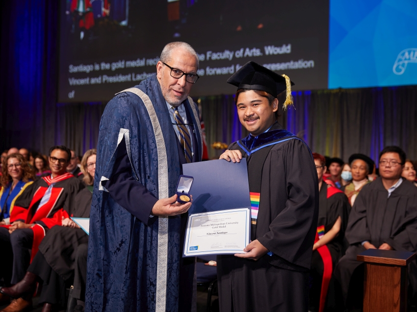 Two people wearing academic regalia pose for a photo while holding a certificate and a gold medal.