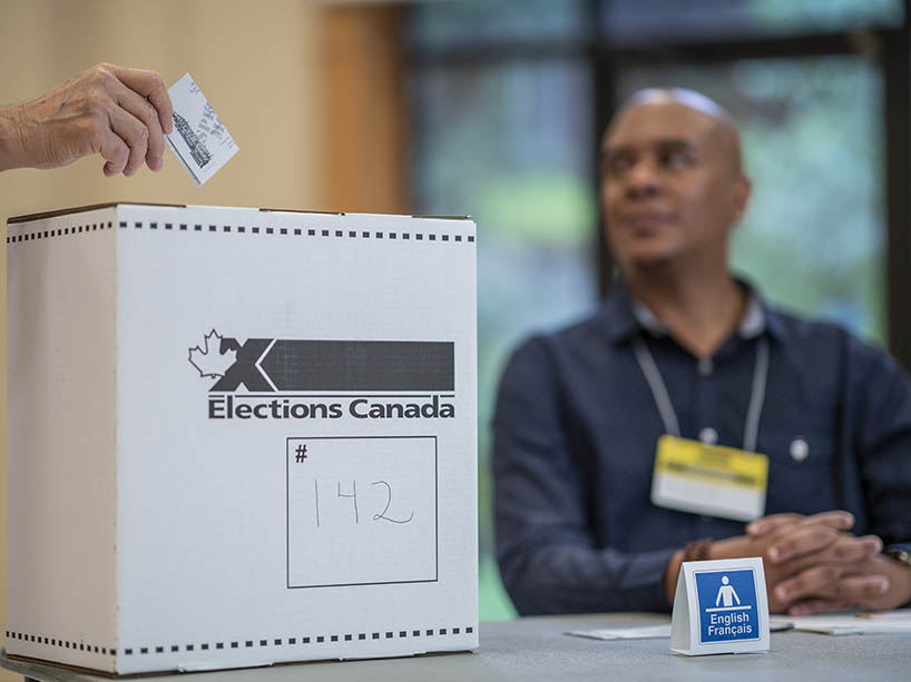 A voter puts a ballot into an Elections Canada ballot box. A polling station worker is in the background.